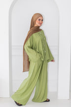 Woman wearing a green outfit with a brown hijab standing against a white wall.