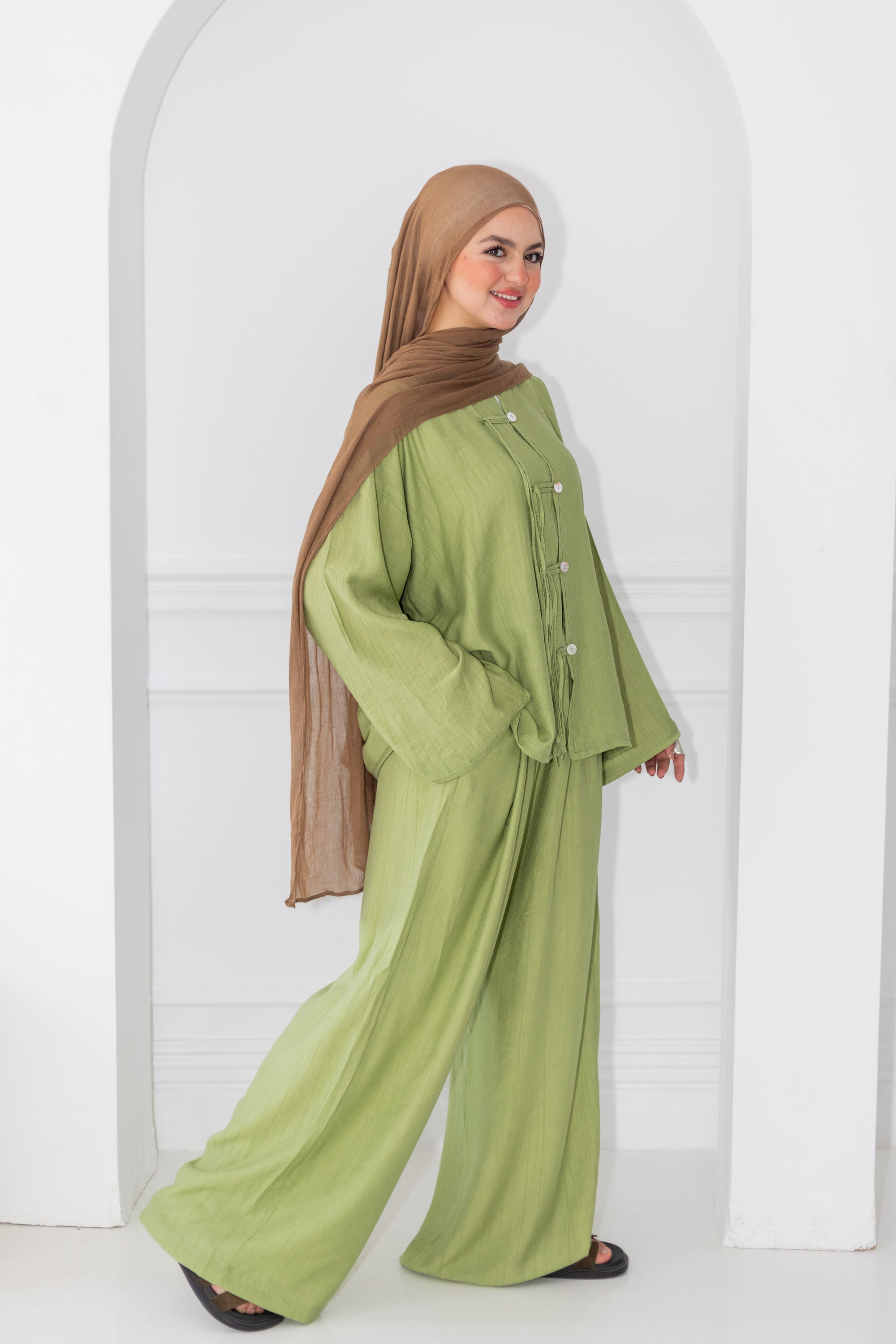 Woman wearing a green outfit with a brown hijab standing against a white wall.