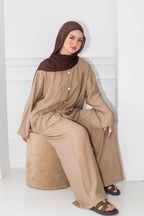 Woman wearing a beige outfit with a brown hijab sitting on a white stool against a white wall.