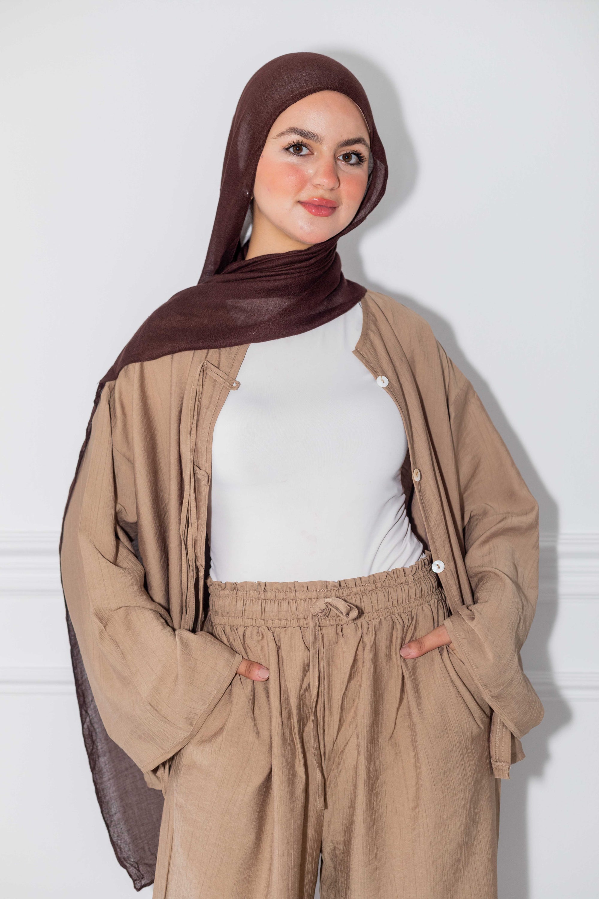 Woman wearing a brown hijab, beige jacket, and pants against a white background