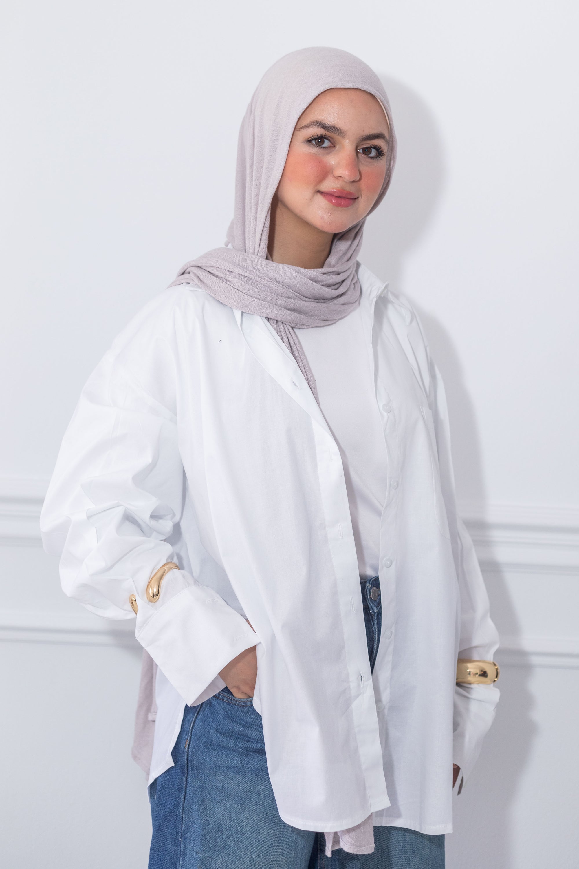 Woman wearing a white shirt and blue jeans with a light gray hijab against a white background