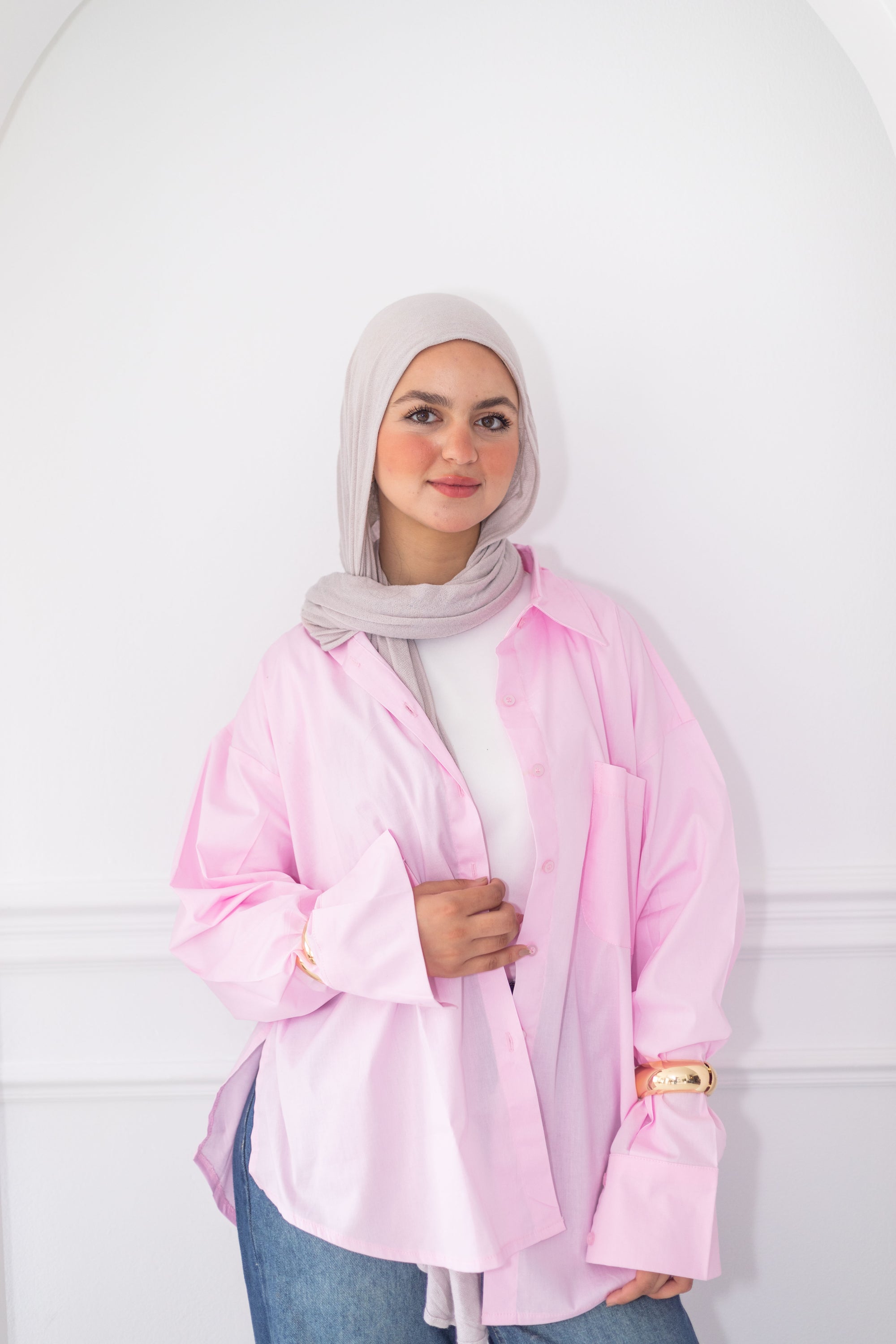 Woman wearing a light pink jacket over a white shirt and blue jeans, standing against a plain background.