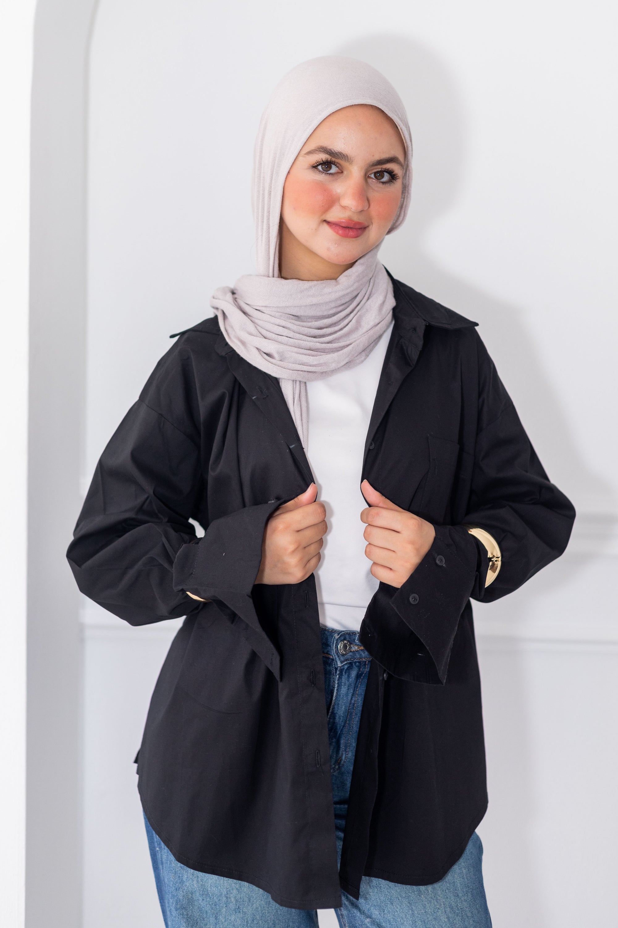 Woman wearing a black jacket, white top, and blue jeans with a light gray hijab against a white background