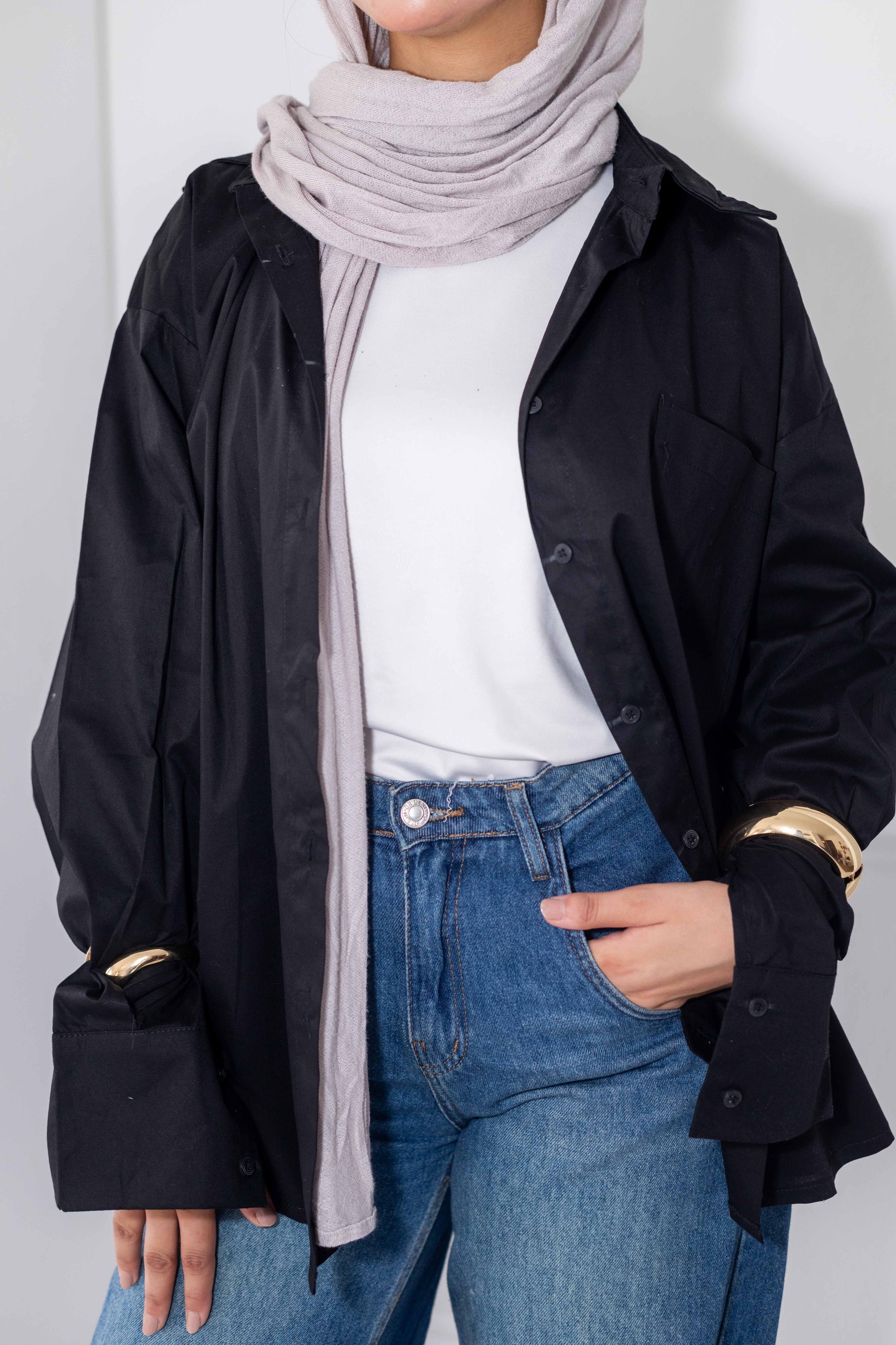 Person wearing a black coat, white shirt, blue jeans, and a light gray hijab against a plain background