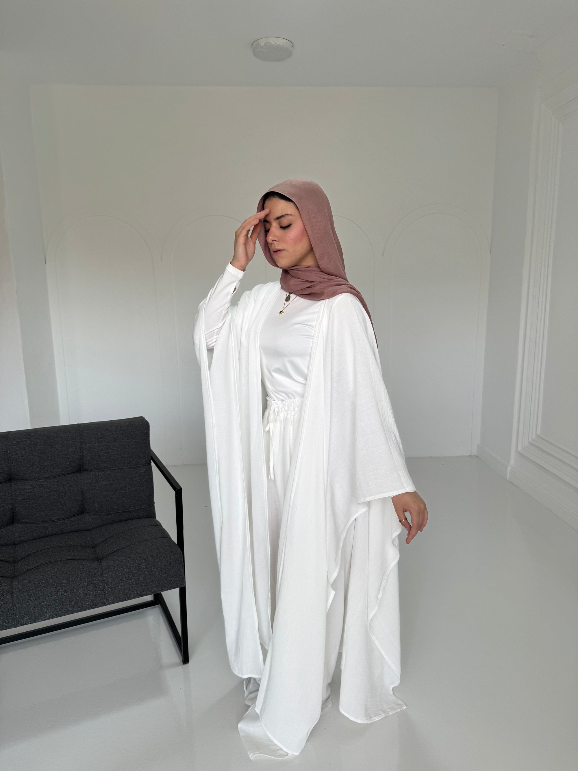 Person wearing a white outfit and brown hijab in a minimalistic room with a black sofa.