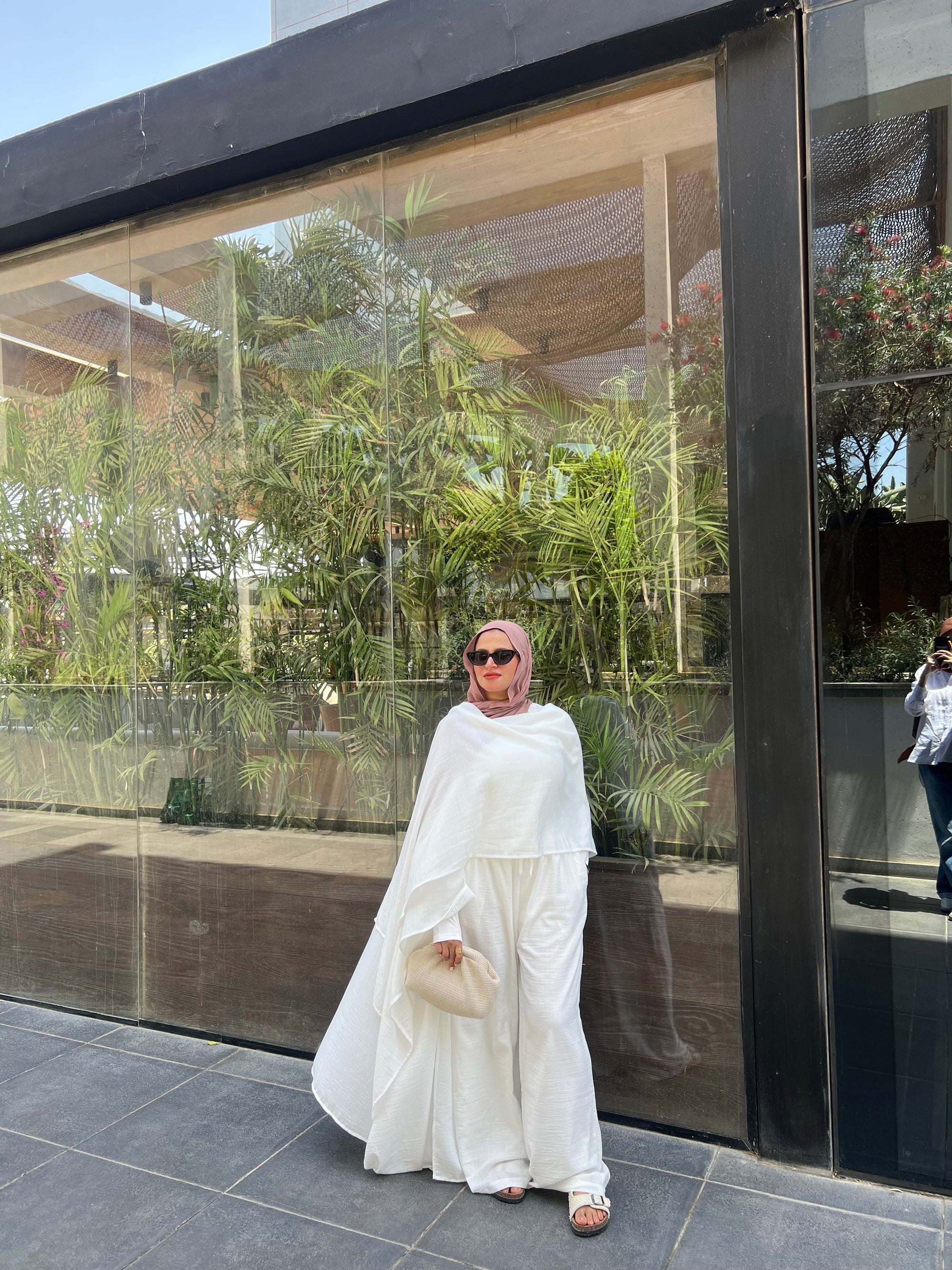 Woman in a white outfit standing in front of a glass wall with greenery outside.