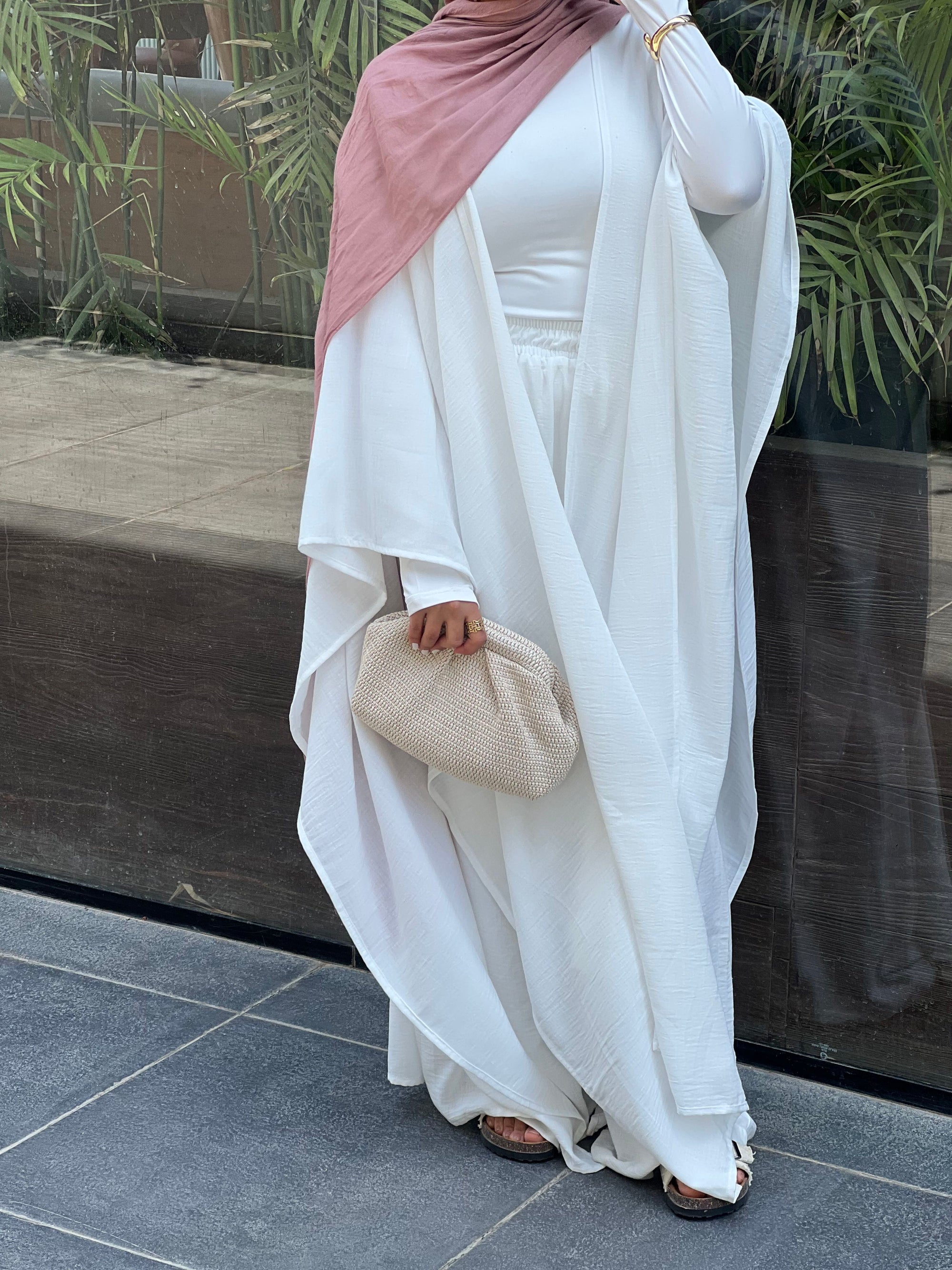 Person wearing a white outfit with a pink headscarf, holding a beige clutch, standing on a tiled floor.