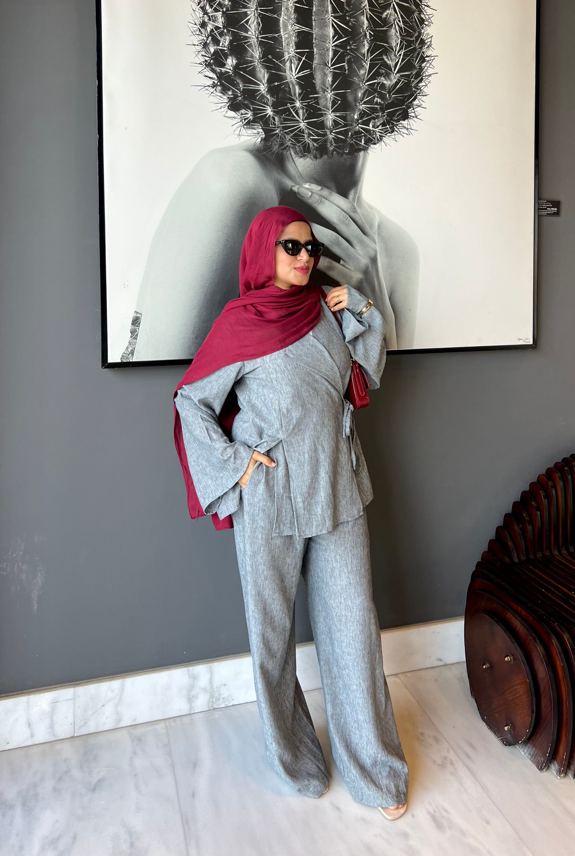 Woman in gray outfit and red hijab standing in front of a wall with a large framed picture.