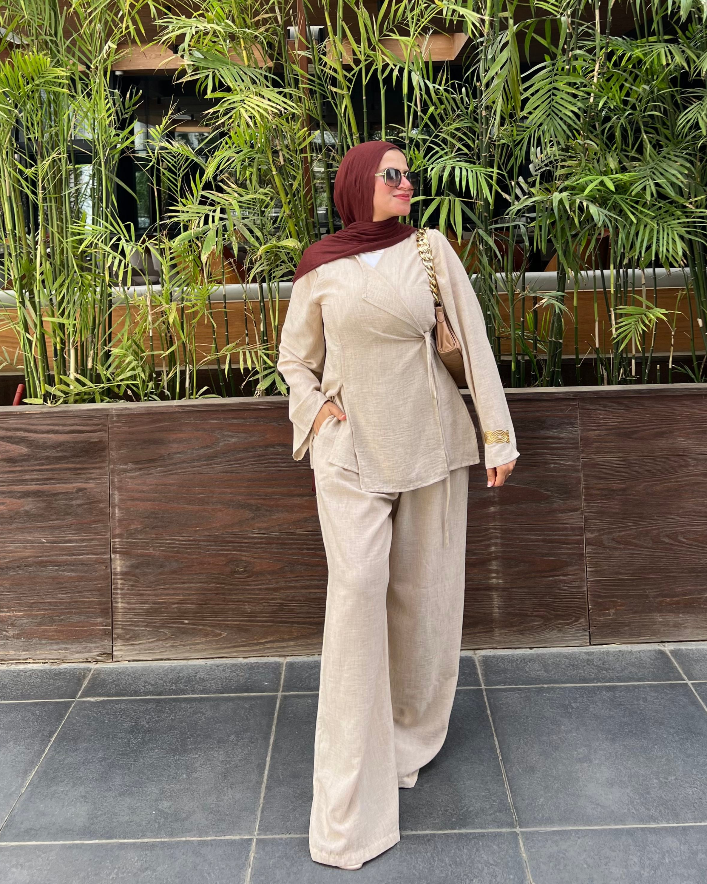 Woman in beige outfit with maroon hijab standing outdoors near plants