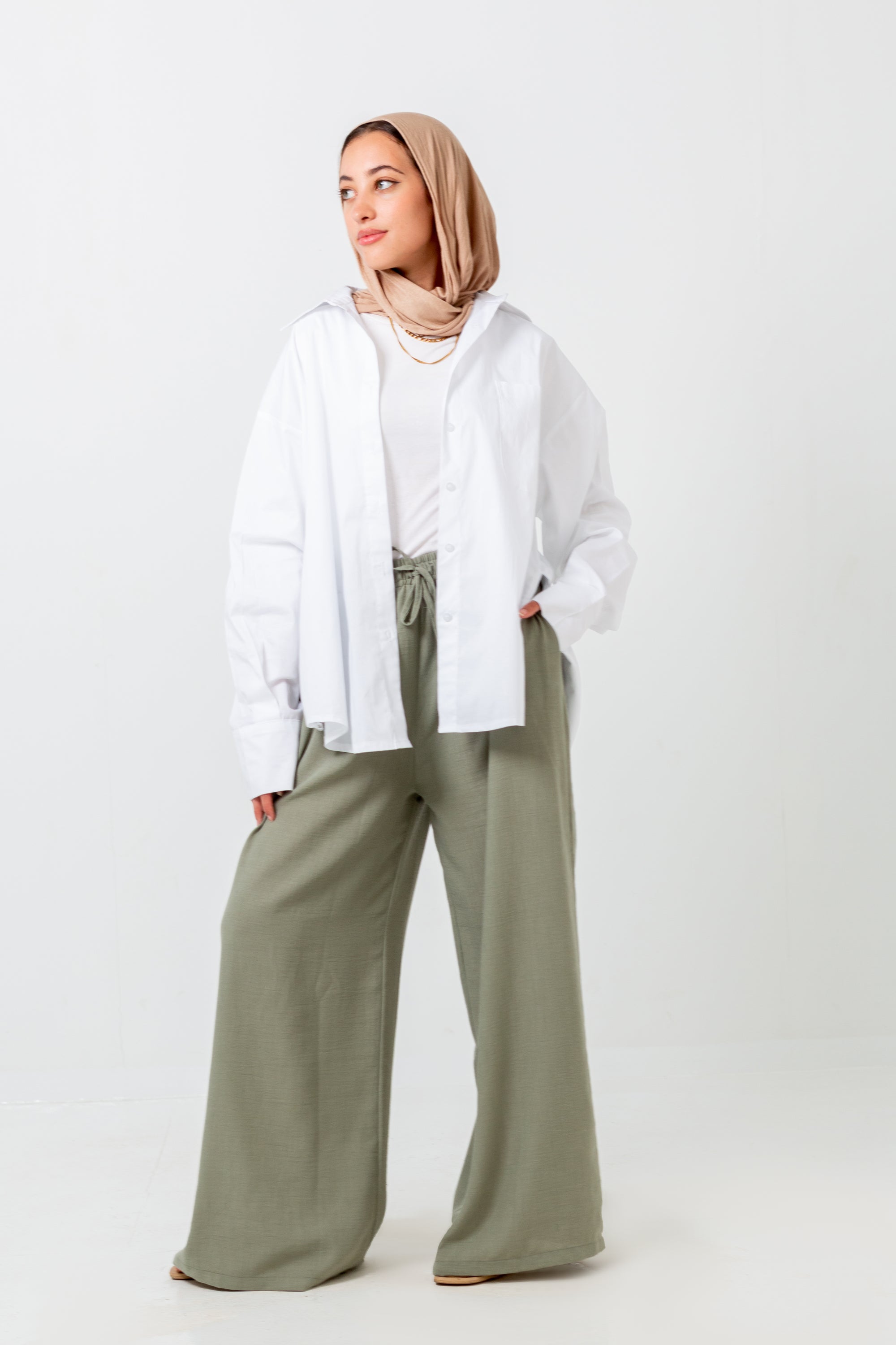 Woman wearing a white shirt, beige hijab, and green pants on a white background