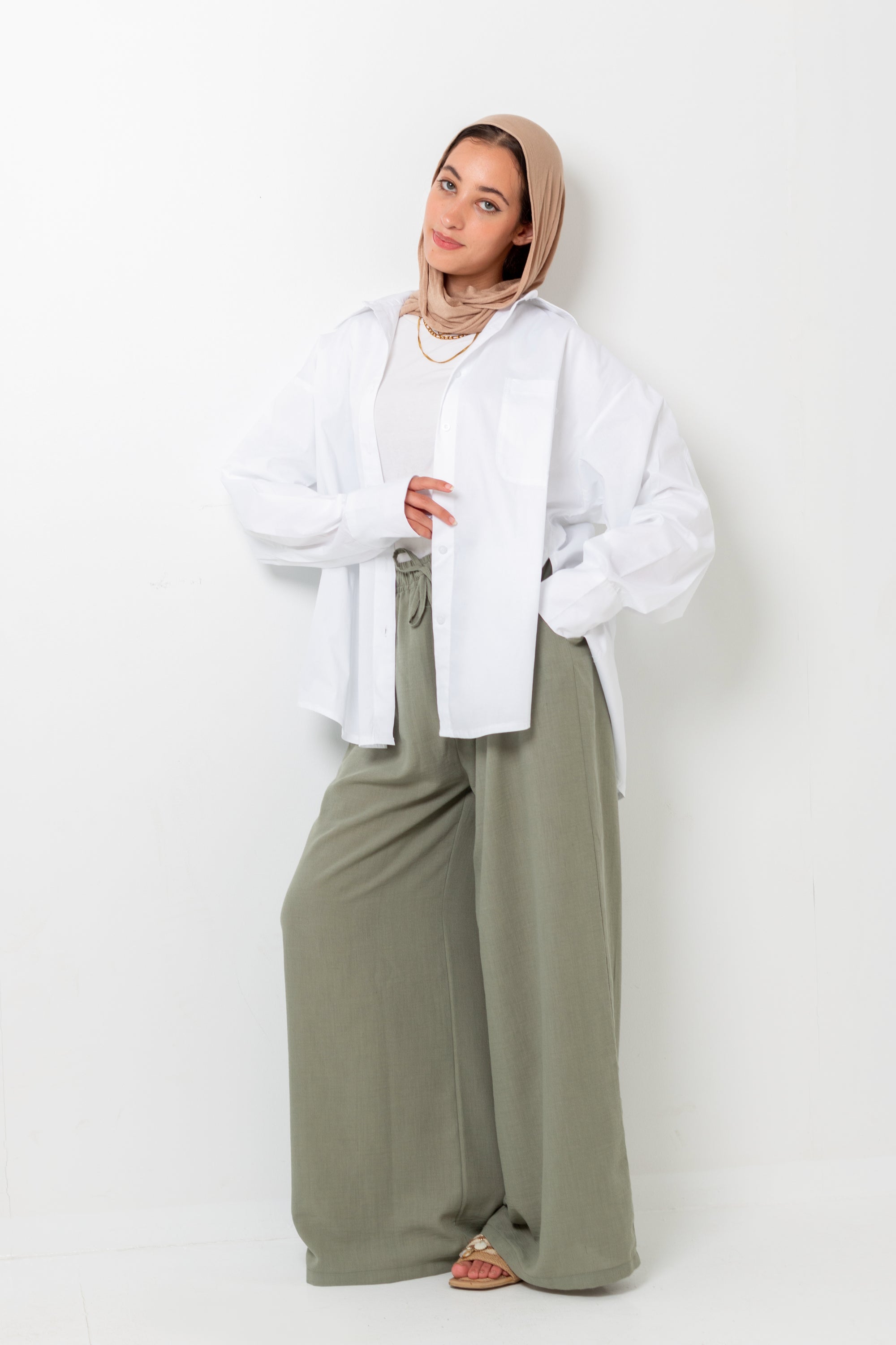 Woman wearing a white shirt and beige hijab with wide-leg green pants on a white background