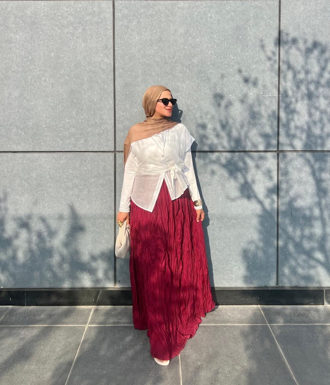 Woman wearing a hijab and a white top with a burgundy skirt against a gray wall.