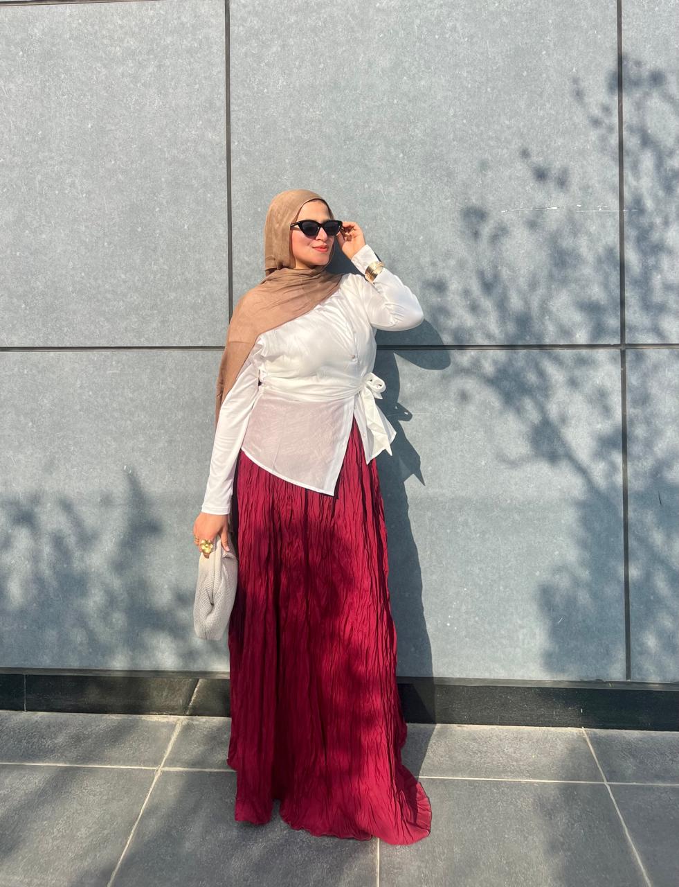 Woman wearing a hijab, white top, and red skirt standing against a gray wall.