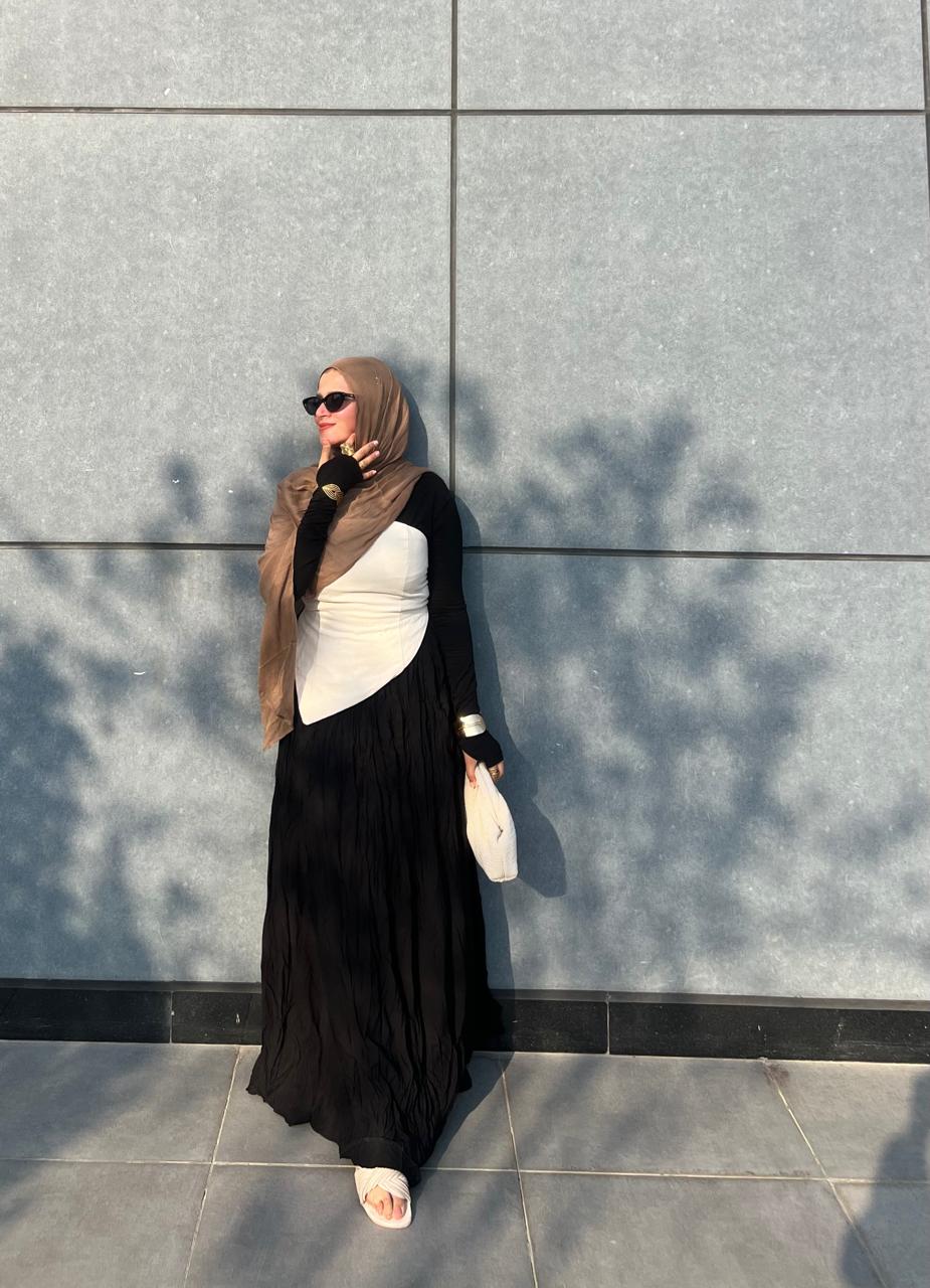 Woman in a hijab and black and white outfit standing against a gray wall.