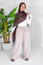 Woman wearing a brown hijab, white top, and beige pants standing next to a plant indoors.