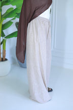 Person wearing a brown top and white pants standing indoors with a plant in the background