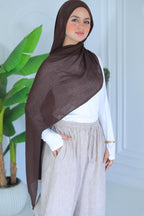 Woman wearing a brown hijab, white top, and beige pants in a room with a plant.