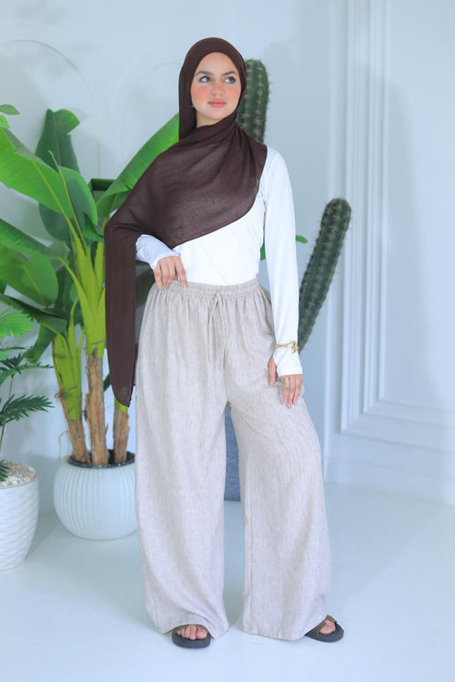 Woman wearing a brown hijab, white top, and beige wide-leg pants standing in a room with plants.