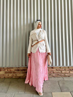 Woman wearing a white top and pink skirt standing against a corrugated metal wall.