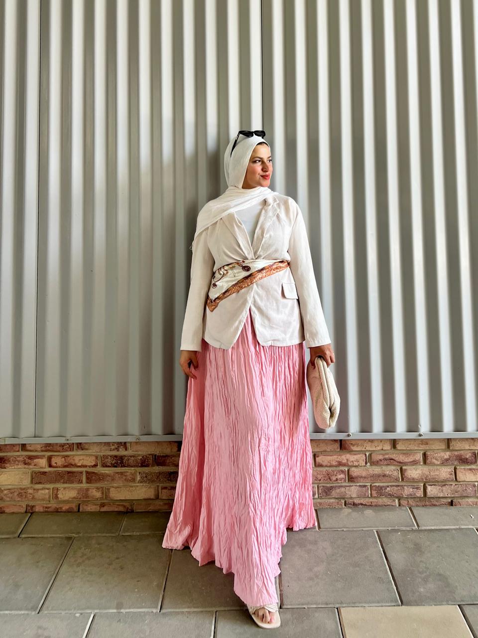 Woman wearing a white top and pink skirt standing against a corrugated metal wall.